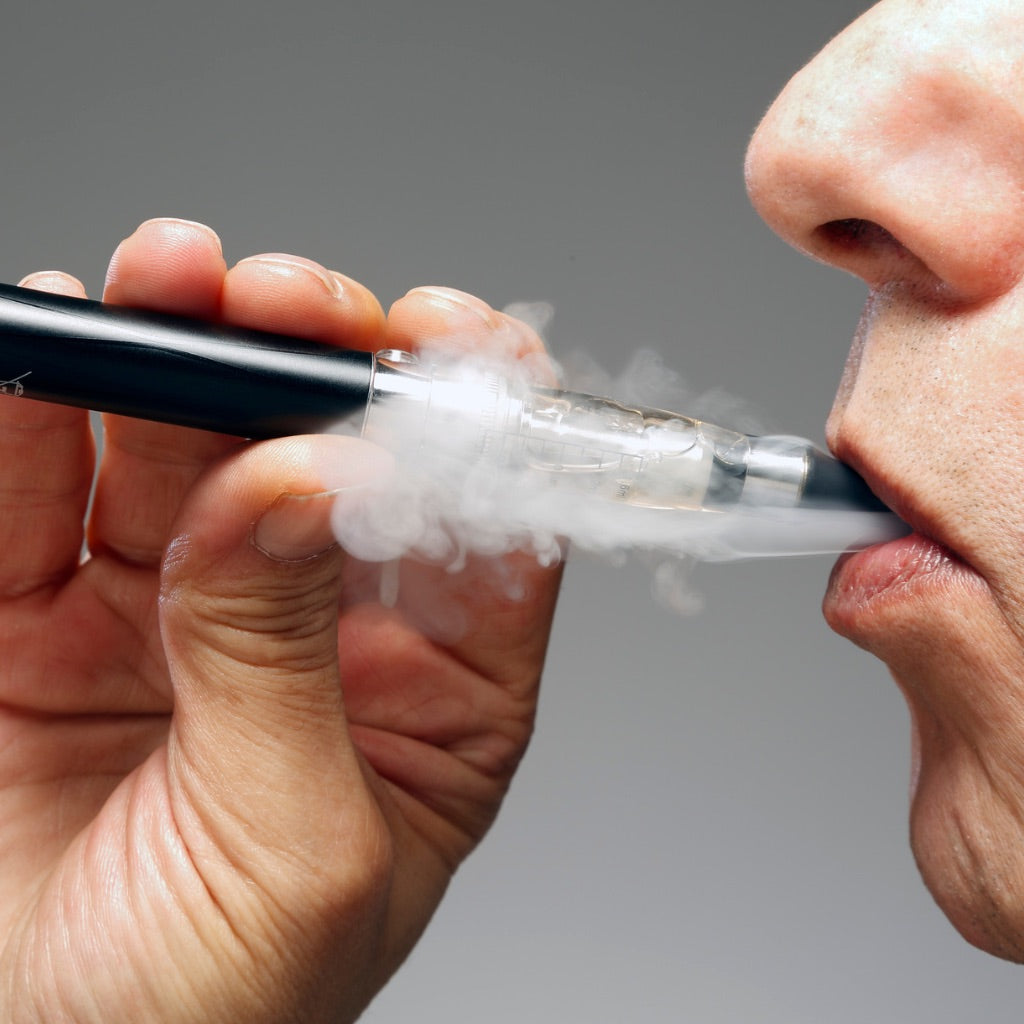 Nahaufnahme Bild von einer Person die eine Vape am Mund hält und den Rauch ausatmet.